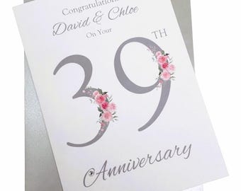 May include: A white anniversary card featuring the text Congratulations David & Chloe On Your 39TH Anniversary. The large grey number 39 is decorated with pink floral illustrations. A small clear crystal accent is placed near the bottom text.