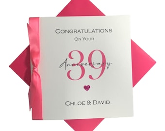 May include: A white anniversary card with the text "Congratulations on your 39th Anniversary" in black and pink. The card is tied with a pink ribbon and set on a pink envelope.