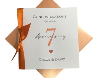 May include: A square white greeting card with a copper-colored ribbon tied on the left side. The text reads CONGRATULATIONS ON YOUR 7 Anniversary with a small copper heart below the number 7 and the names CHLOE & DAVID at the bottom.