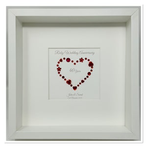 Può includere: Un'opera d'arte incorniciata che celebra un anniversario di matrimonio di rubino. La cornice bianca circonda uno sfondo bianco con un disegno a forma di cuore fatto di gemme rosse. Il testo recita "Ruby Wedding Anniversary" e "40 Years".