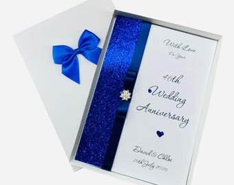 May include: A white anniversary card with a blue satin bow on the lid. The card inside has a blue glitter strip and a blue ribbon with a jewel. Text reads "With Love On Your 46th Wedding Anniversary" and the names "David & Chloe" with the date "25th July 2025."