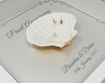 May include: A framed pearl anniversary keepsake. The white frame surrounds a light gray background with a white seashell holding a single pearl. Text reads "Pearl Anniversary" and names with a date.