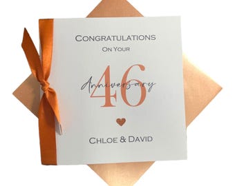 May include: A white anniversary card with the text "Congratulations on your 46 Anniversary" in black and coral. The card is tied with an orange ribbon and sits on a coral envelope. The names "Chloe & David" are printed at the bottom.