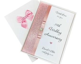 May include: A white gift box with a pink satin bow on the lid. Inside, a card with a pink glitter strip and a ribbon with a jewel. The card reads "39th Wedding Anniversary" with names and a date.
