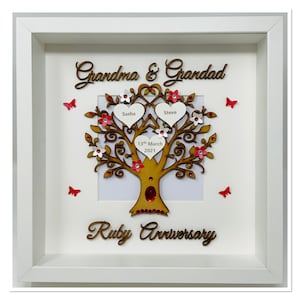 Può includere: Una cornice bianca con un albero genealogico in legno. Contiene le parole "Grandma & Grandad" e "Ruby Anniversary". L'albero ha foglie a forma di cuore con nomi e una data, adornato con fiori e farfalle rosse e bianche.
