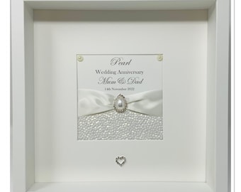 May include: A framed pearl wedding anniversary gift. The white frame surrounds a white background with text that reads "Pearl Wedding Anniversary Mum & Dad 14th November 2022". A white ribbon with a pear-shaped jewel is centered.