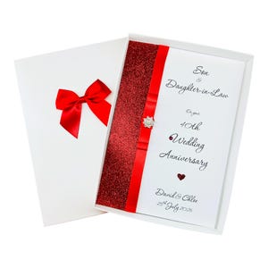 Pode incluir: Uma caixa de presente branca contendo um cartão de felicitações para um 40º aniversário de casamento. O cartão apresenta um painel de glitter vermelho, uma fita de cetim vermelha com um enfeite de cristal e o texto Son & Daughter-in-Law, David & Chloe, 25th July 2026.