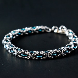 Puede incluir: Un brazalete de malla de cadena plateado y azul turquesa. El brazalete está hecho de pequeños anillos de metal que están entrelazados para crear un patrón de malla de cadena.