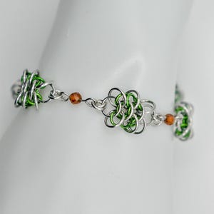 Könnte beinhalten: Ein grünes und silbernes Kettenarmband mit braunen Perlen. Das Armband besteht aus kleinen Metallringen, die miteinander verbunden sind, um eine Kette zu bilden. Die Kette ist mit kleinen, runden Perlen verziert, die eine dunkelbraune Farbe haben.