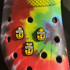 May include: Three yellow and black US Navy charm shoe charms with an anchor and the letters USN. The charms are on a tie-dye background.