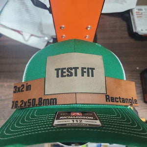 May include: Green and white trucker hat with a brown patch that says "TEST FIT" and "3x2 in 76.2x50.8mm Rectangle". The hat has a Richardson 112 adjustable label.