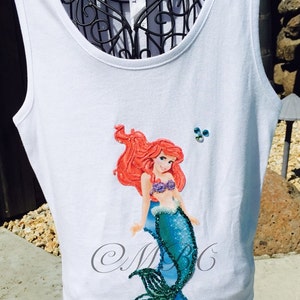 May include: White tank top with a red-haired mermaid applique with a blue tail and green glitter accents. The mermaid is wearing a purple seashell bra. The tank top has the text "MBG" in cursive on the bottom.