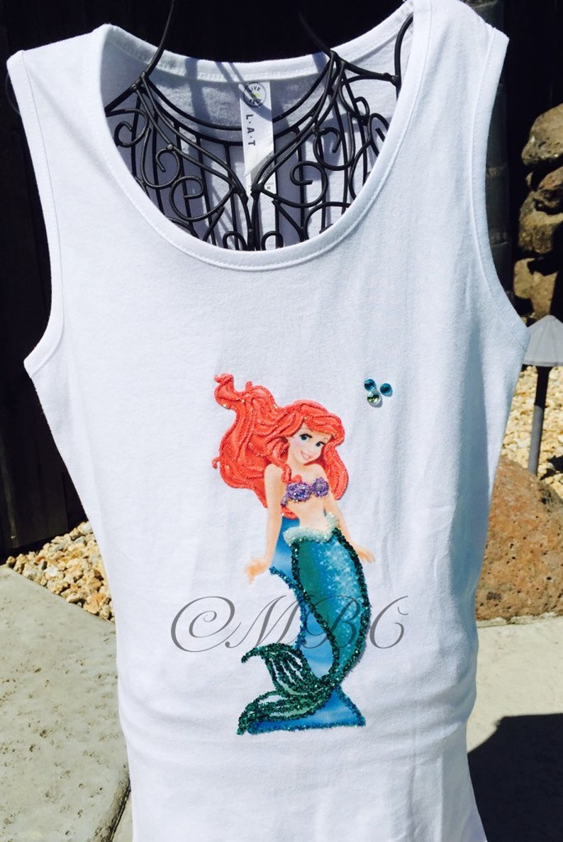 May include: White tank top with a red-haired mermaid applique with a blue tail and green glitter accents. The mermaid is wearing a purple seashell bra. The tank top has the text "MBG" in cursive on the bottom.