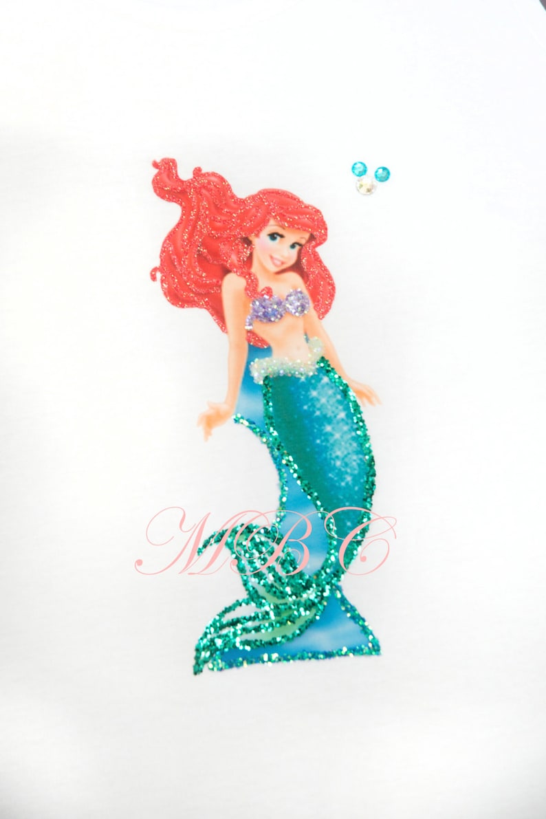 May include: A glittery mermaid applique with red hair and a turquoise tail. The applique is on a white background with two small turquoise rhinestones.