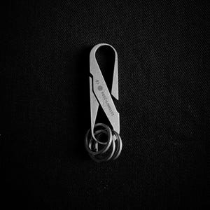 Può includere: Un portachiavi argentato con un design curvo e un set di portachiavi. Il portachiavi ha incisa la scritta "#3 KEYCA/ARRIVE". Lo sfondo è nero pieno.