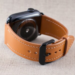 May include: A tan leather watch strap with a black buckle and stitching. The strap is designed for an Apple Watch.