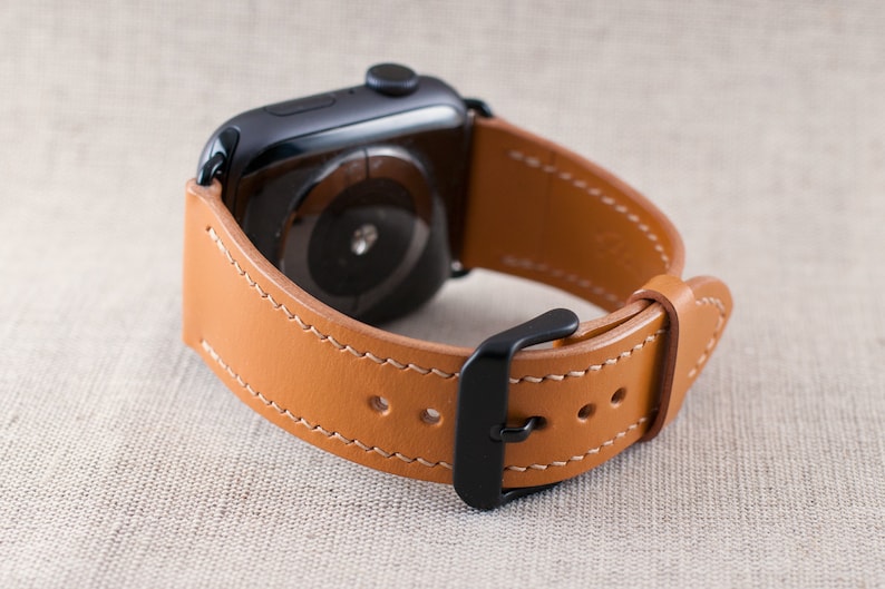 May include: A tan leather watch strap with a black buckle and stitching. The strap is designed for an Apple Watch.