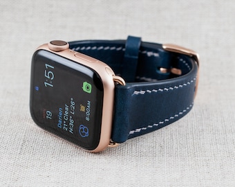 May include: A rose gold Apple Watch with a dark blue leather strap. The watch face shows the time, date, and weather information. The strap has white stitching.