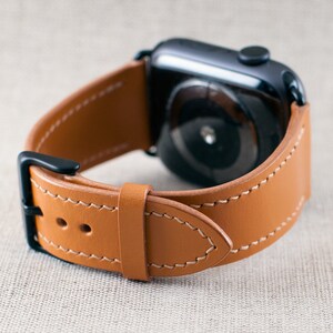 May include: A brown leather watch strap with white stitching. The strap is designed for an Apple Watch and has a black metal buckle.