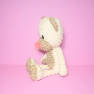 May include: A crocheted teddy bear with a light brown body and a cream-coloured head. The bear has a pink nose and black eyes.