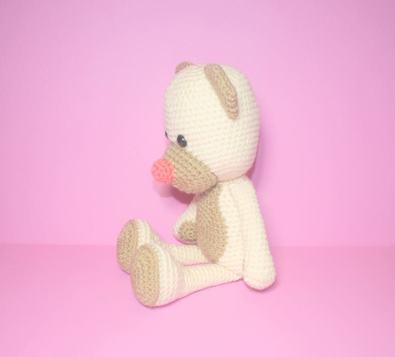 May include: A crocheted teddy bear with a light brown body and a cream-coloured head. The bear has a pink nose and black eyes.