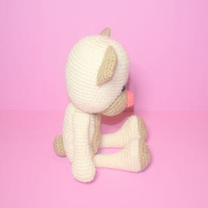 May include: A crocheted stuffed animal in the shape of a bear. The bear is sitting with its legs crossed and has a pink nose. The bear is white with brown ears and is sitting on a pink background.
