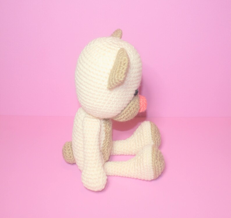 May include: A crocheted stuffed animal in the shape of a bear. The bear is sitting with its legs crossed and has a pink nose. The bear is white with brown ears and is sitting on a pink background.