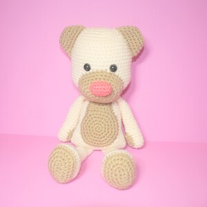May include: A crocheted stuffed animal dog with brown and white fur, a pink nose, and black eyes. The dog is sitting on a pink background.