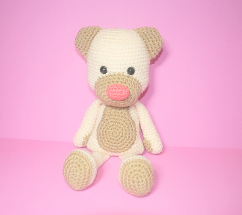 May include: A crocheted stuffed animal dog with brown and white fur, a pink nose, and black eyes. The dog is sitting on a pink background.