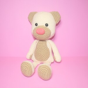 May include: A crocheted stuffed toy in the shape of a dog. The dog is beige and white with brown eyes and a pink nose. It is sitting on a pink background.