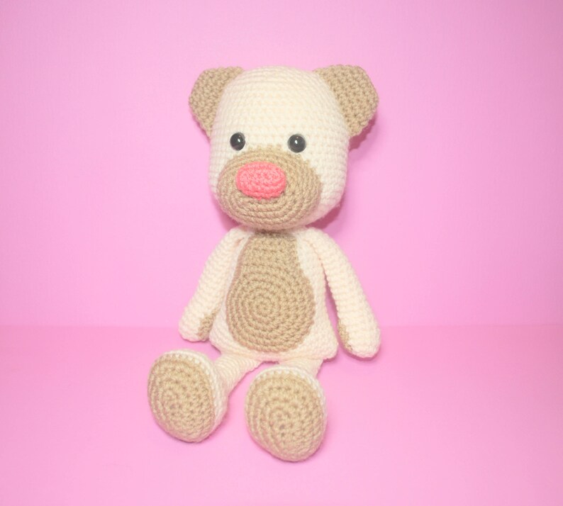 May include: A crocheted stuffed toy in the shape of a dog. The dog is beige and white with brown eyes and a pink nose. It is sitting on a pink background.
