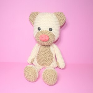 May include: A crocheted stuffed animal in the shape of a bear. The bear is cream coloured with brown accents and has a pink nose. The bear is sitting on a pink background.