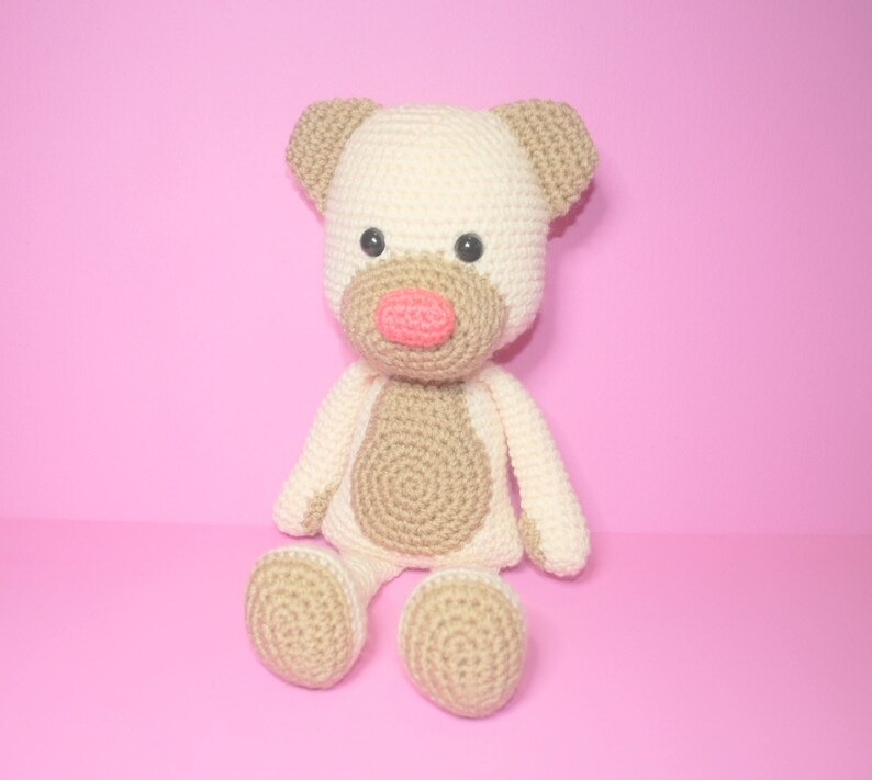 May include: A crocheted stuffed animal in the shape of a bear. The bear is cream coloured with brown accents and has a pink nose. The bear is sitting on a pink background.