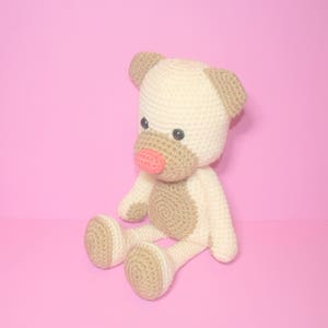 May include: A crocheted stuffed toy in the shape of a dog. The dog is sitting with its legs crossed and has a light brown body with a cream-coloured head and ears. The dog has a pink nose and black eyes.