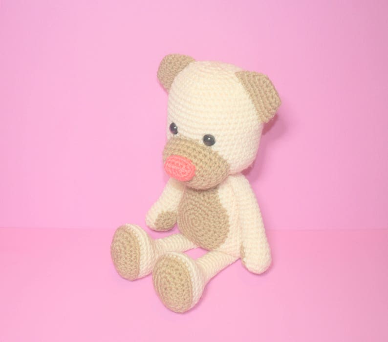 May include: A crocheted stuffed toy in the shape of a dog. The dog is sitting with its legs crossed and has a light brown body with a cream-coloured head and ears. The dog has a pink nose and black eyes.