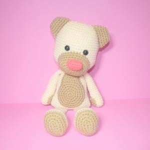 May include: A crocheted stuffed toy in the shape of a dog. The dog is beige with brown accents and has a pink nose. The dog is sitting on a pink background.