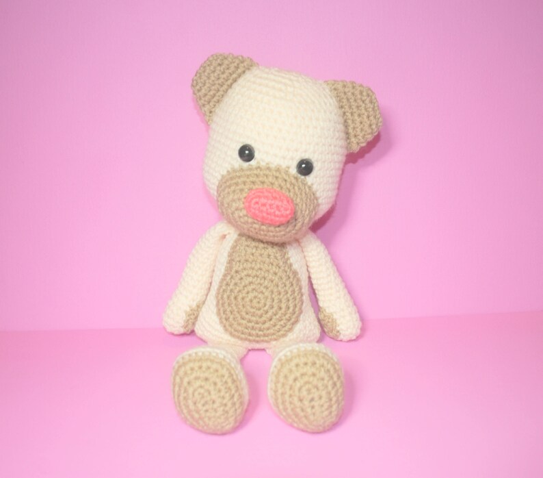 May include: A crocheted stuffed toy in the shape of a dog. The dog is beige with brown accents and has a pink nose. The dog is sitting on a pink background.
