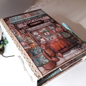 Puede incluir: Un álbum de recortes azul y blanco con una imagen de fachada de tienda vintage en la portada. La fachada tiene un letrero que dice "Brocante Antiques". Hay un oso de peluche sentado frente a la tienda.