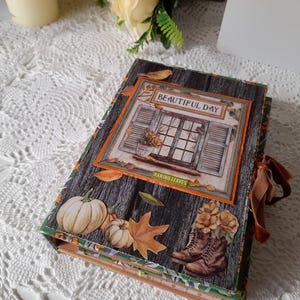 Puede incluir: Un álbum de recortes rectangular con una cubierta de madera oscura que presenta imágenes otoñales, incluyendo calabazas, hojas caídas y un par de botas marrones. Una ilustración central muestra una ventana con el texto BEAUTIFUL DAY y RAKING LEAVES.