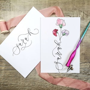 May include: Two white rectangular cards featuring the name Sarah written in black cursive calligraphy. One card includes a watercolor illustration of three sweet pea flowers in pink, red, and light purple. A pink pen and a spool of pink ribbon are present.
