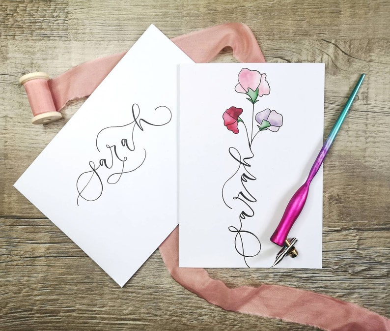 May include: Two white rectangular cards featuring the name Sarah written in black cursive calligraphy. One card includes a watercolor illustration of three sweet pea flowers in pink, red, and light purple. A pink pen and a spool of pink ribbon are present.