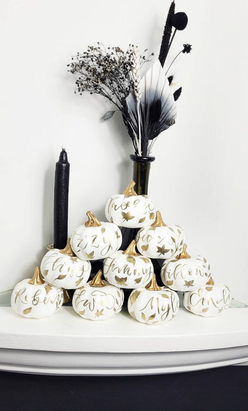 Pu&ograve; includere: Una collezione di piccole zucche bianche con piccioli dorati, dipinte a mano con nomi in calligrafia dorata e motivi a foglia. Le zucche sono impilate a piramide, accompagnate da una candela nera e da una composizione di fiori secchi scuri.