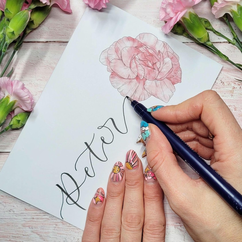 May include: A white sheet of paper featuring a hand-drawn pink carnation illustration and the name Peter written in black cursive calligraphy. A dark blue pen is positioned on the paper, held by fingers with colorful patterned nail art.