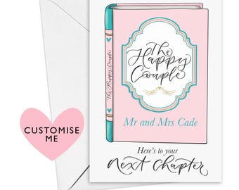 Pode incluir: Um cartão rosa e azul em forma de livro com o texto "The Happy Couple" e "Mr and Mrs Cade". O cartão também tem o texto "Here's to your Next Chapter".