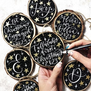 Può includere: Ornamenti natalizi in legno dipinti a mano con sfondi neri, stelle dorate e calligrafia bianca. Gli ornamenti presentano frasi come "Joy", "1st Christmas Engaged 2023" e nomi con l'anno 2023. Ogni ornamento ha un anello di spago.