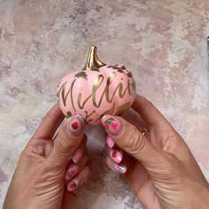 Pu&ograve; includere: Una piccola zucca decorativa rotonda di colore rosa chiaro, dotata di un picciolo color oro metallizzato e della scritta Millie in corsivo dorato. La superficie &egrave; decorata con motivi a foglia d'oro e piccoli disegni floreali.