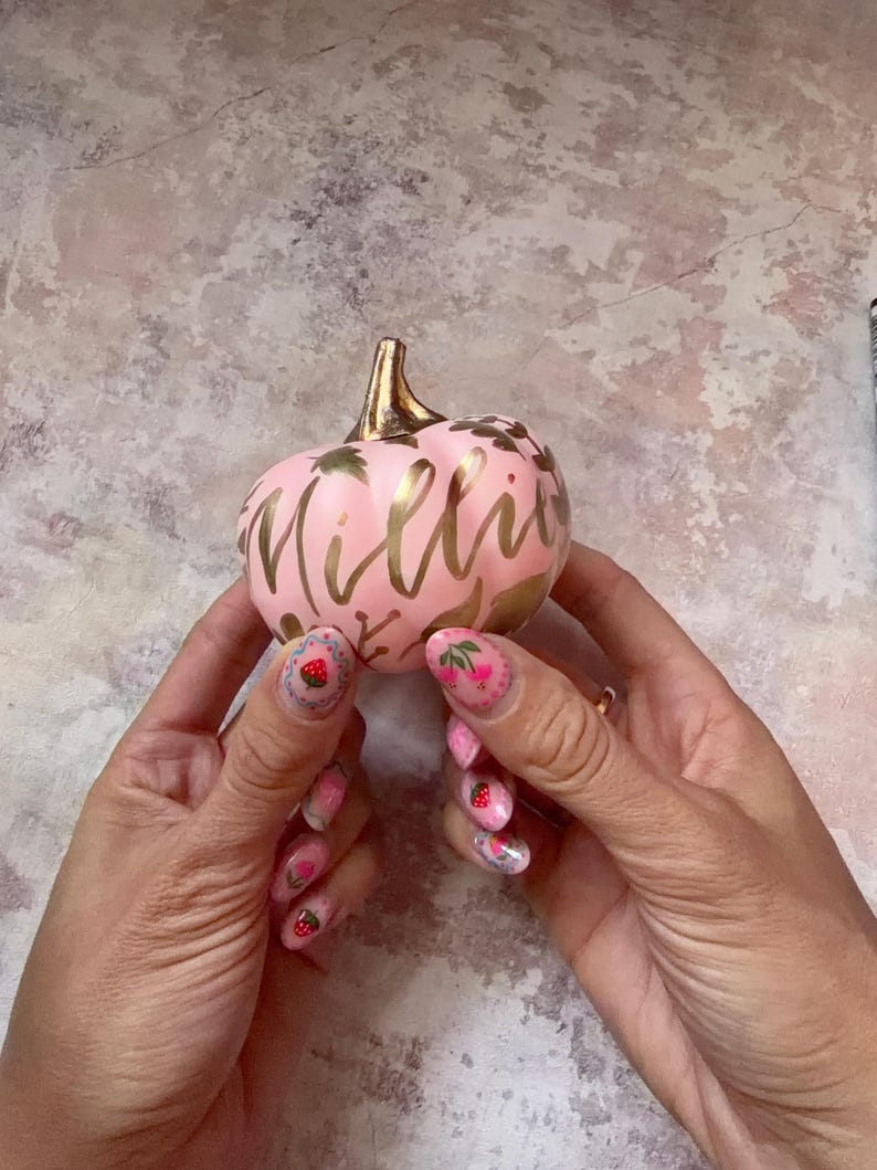 Pu&ograve; includere: Una piccola zucca decorativa rotonda di colore rosa chiaro, dotata di un picciolo color oro metallizzato e della scritta Millie in corsivo dorato. La superficie &egrave; decorata con motivi a foglia d'oro e piccoli disegni floreali.
