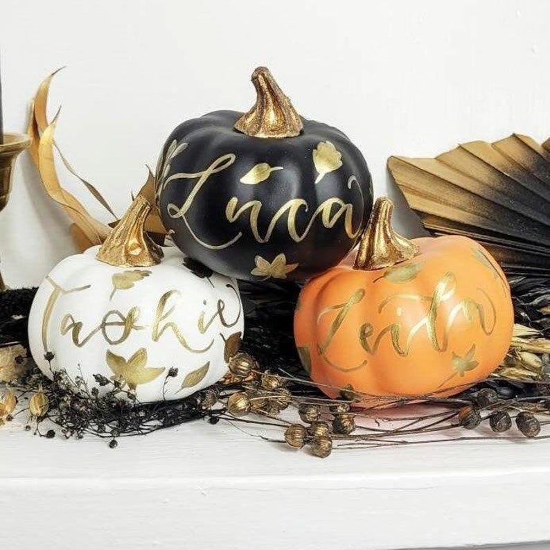 Pu&ograve; includere: Tre zucche decorative in bianco, nero e arancione, ognuna con un picciolo dorato metallizzato e nomi e motivi a foglia dipinti a mano in calligrafia dorata. La zucca bianca riporta "Archie", quella nera "Luca" e quella arancione "Zeith".