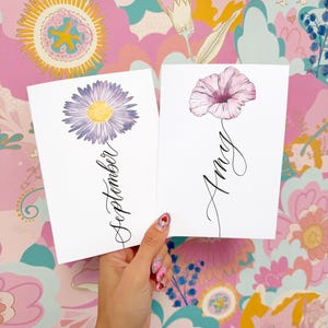 May include: Two white rectangular cards featuring floral illustrations. The left card displays a purple daisy with a yellow center and the word September in black script. The right card shows a pink flower with the name Amy in black script.