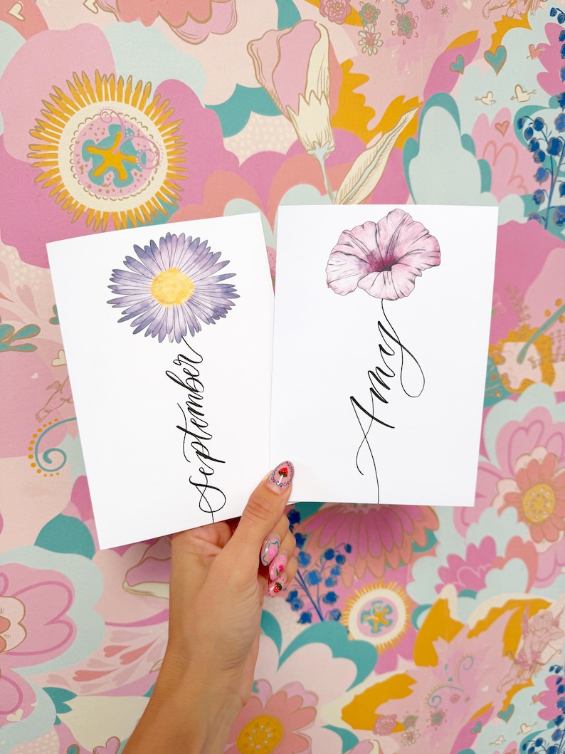 May include: Two white rectangular cards featuring floral illustrations. The left card displays a purple daisy with a yellow center and the word September in black script. The right card shows a pink flower with the name Amy in black script.